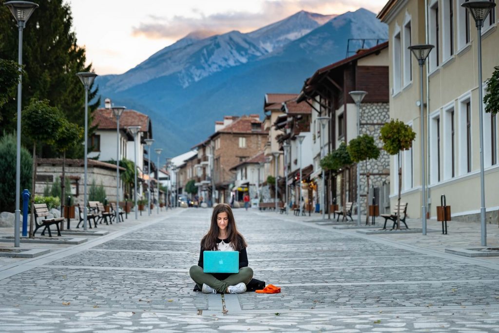 An image featuring a woman, a digital nomad, sitting in the middle of a road, engrossed in her work on a laptop, showcasing the freedom and flexibility of remote work and the ability to work from anywhere.