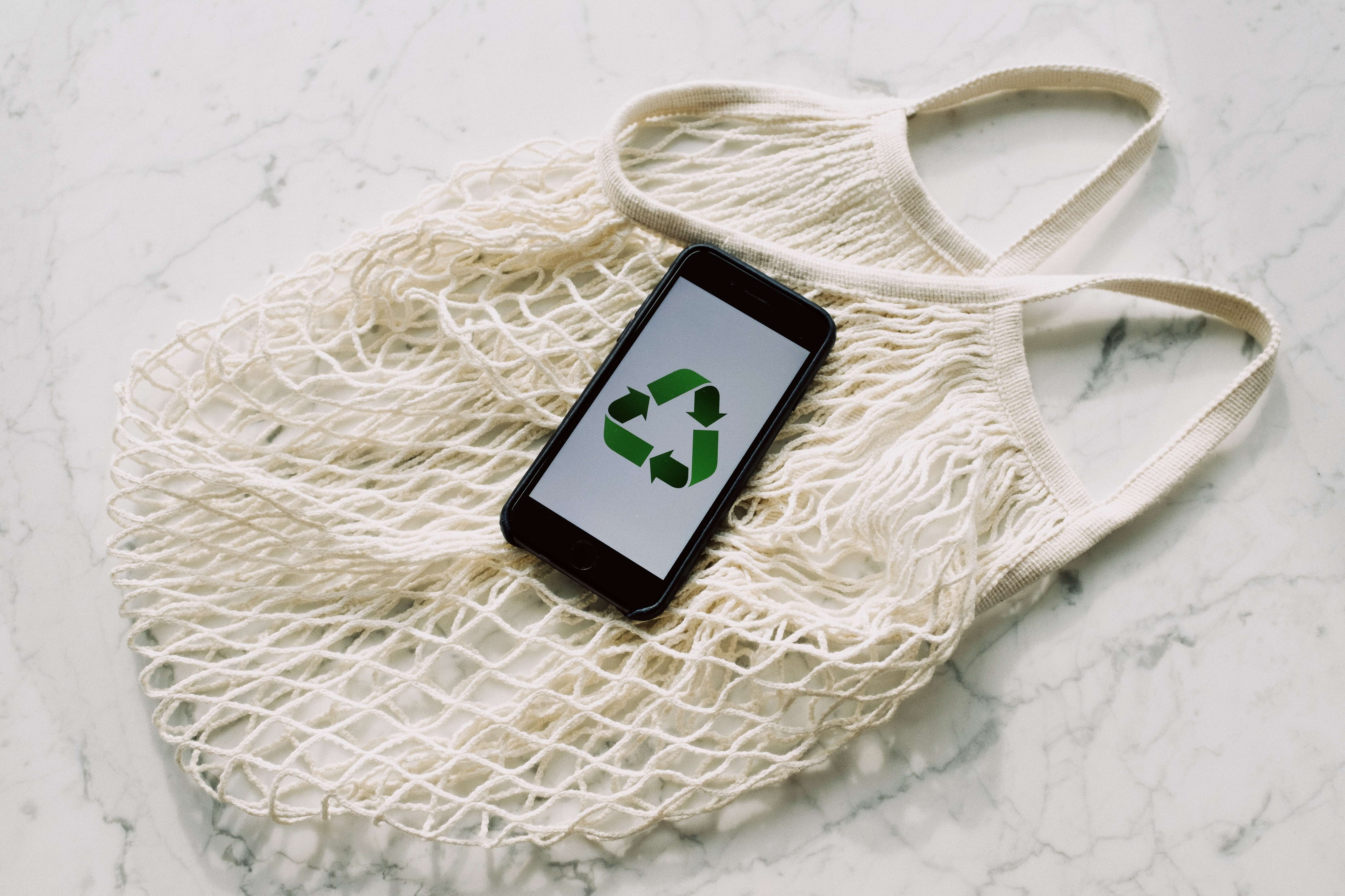 An image featuring a cloth bag with a phone resting on top of it, accompanied by the universally recognized symbol for recycling, representing the concept of environmentally conscious practices and the integration of technology in sustainable living.